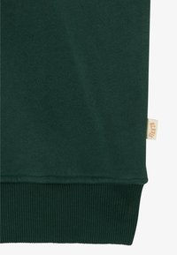 Dunkelgrüner Stoff mit einem gerippten Saum und einem kleinen beigen Etikett mit der Aufschrift "BLKMIS", das an der Seitennaht angenäht ist.