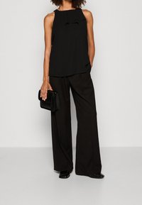 Schwarzes ärmelloses Top mit Rüschendetails, kombiniert mit lockeren schwarzen Hosen. Das Model hält eine schwarze strukturierte Clutch und trägt schwarze Schuhe.