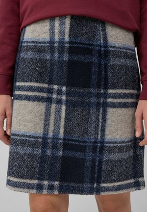 Frau in einem karierten Wollrock in Blau, Schwarz und Beige, kombiniert mit einem burgunderroten Langarmoberteil, gezeigt von der Taille bis zu den Knien.