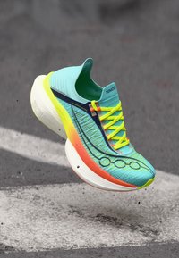 Αθλητικό παπούτσι γαλάζιου και νέον κίτρινου χρώματος με πορτοκαλί λεπτομέρειες, αιωρούμενο πάνω από άσφαλτο με φθαρμένες λευκές γραμμές.