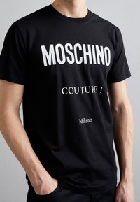 Man med svart t-shirt med vit text "MOSCHINO," "COUTURE!" och "Milano," står med en hand i fickan mot en enfärgad bakgrund.