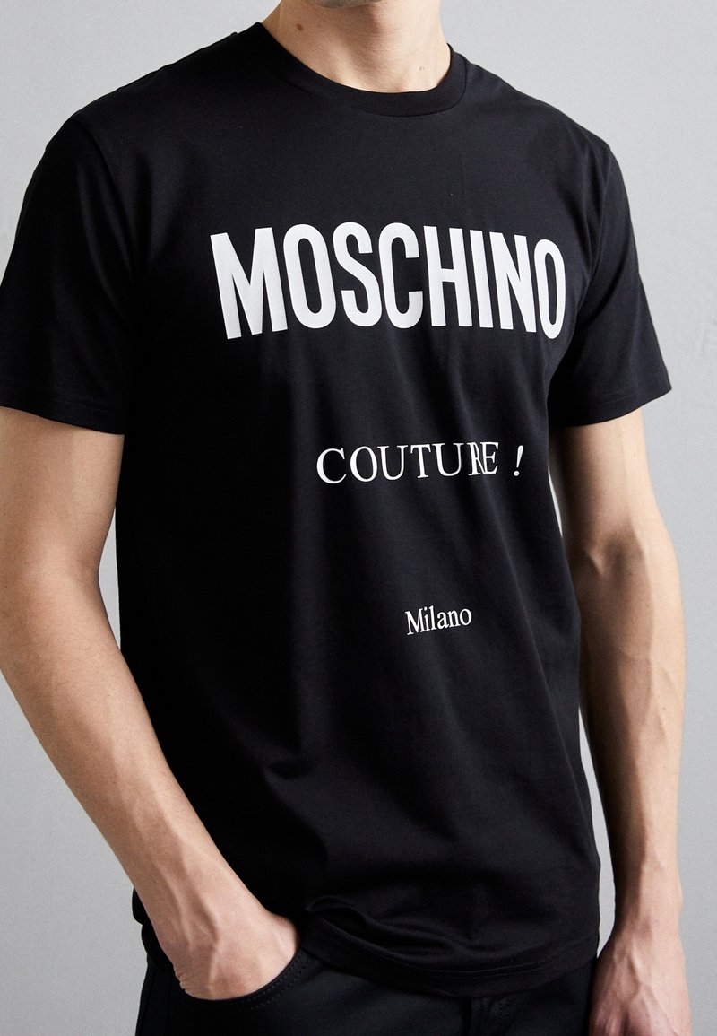 Man med svart t-shirt med vit text "MOSCHINO," "COUTURE!" och "Milano," står med en hand i fickan mot en enfärgad bakgrund.