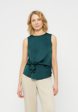 Femeie cu păr scurt blond, purtând o bluză verde închis fără mâneci, cu nod în față, și pantaloni bej deschis, stând în picioare pe un fundal alb.