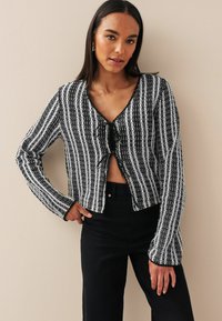Cardigan accorciato a righe bianche e nere con maniche lunghe, scollatura a V e dettaglio di nodo frontale. Tessuto a maglia testurizzato con bordi scallop.