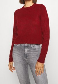 Pull en tricot rouge avec un col ras du cou, des poignets et un ourlet côtelés. Associé à un jean ajusté gris clair. Textured lisse et design décontracté.