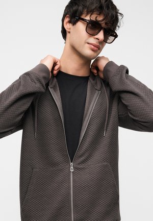 Hnedá mikina na zips s textúrovaným prešívaným vzorom, klokanie vrecká a kapucňou so sťahovacou šnúrkou, v kombinácii s čiernym tričkom pod ňou.