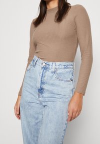 Una maglietta a maniche lunghe a coste marrone chiaro abbinata a jeans in denim azzurro chiaro a vita alta. I jeans sono sbiaditi con cuciture e tasche ben visibili.