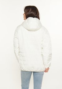 Hvid pufferjakke med hætte, der har et quiltet mønster, elastisk kant og strikkede manchetter. Båret over lyseblå jeans.