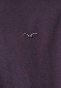 Dark purple t-shirt made of cotton. Features a small, light purple embroidered seagull logo near the center. Soft, smooth texture.