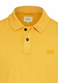 Żółta koszulka polo wykonana z teksturowanej tkaniny, z kołnierzykiem, posiadająca czarne guziki oraz małe haftowane logo po lewej stronie klatki piersiowej.
