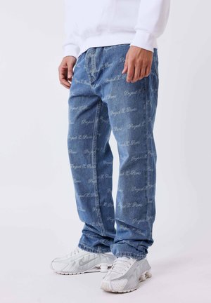 Lichtblauwe denimjeans met herhaalde witte tekstpatronen, een losse pasvorm en zichtbare stiksels. Gepaarde met witte sportschoenen.