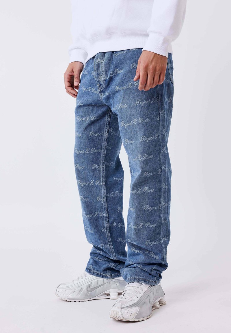 Los jeans de mezclilla de color azul claro presentan patrones de texto blanco repetidos, un corte holgado y costuras visibles. Se combinan con zapatillas deportivas blancas.