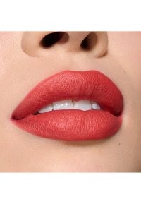 Matte rød læbestift på fyldige læber, som viser en glat, fløjlsagtig tekstur. Hvide tænder er synlige, med en neutral baggrund.
