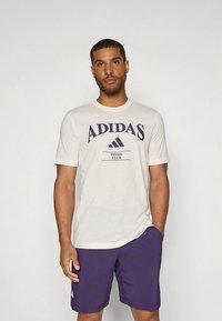Camiseta Adidas de manga corta color crema con letras moradas que dicen "ADIDAS" y "TENNIS CLUB", combinada con pantalones cortos morados. Diseño sencillo, tela suave.