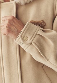 Beżowy wełniany płaszcz z miękkim kołnierzem i haftem misia. Zawiera mankiet na guziki z ozdobnym metalowym guzikiem oraz wyprofilowane rękawy.