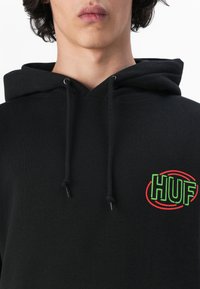 Black hoodie with dual drawstrings, featuring a green and red "HUF" logo on the left chest. Made of soft fabric, with a relaxed fit.