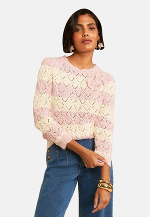 Vrouw draagt een roze en crème gestreepte gebreide trui met bloemdetails en blauwe high-waist jeans, geaccentueerd met grote gouden armbanden.