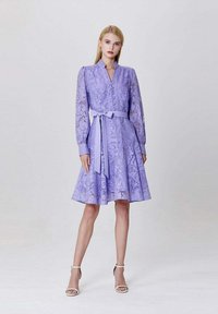 Robe violette à manches longues, motif floral, avec un devant boutonné et une ceinture à la taille. Jupe évasée et arrivant au genou. Associée à des sandales blanches.