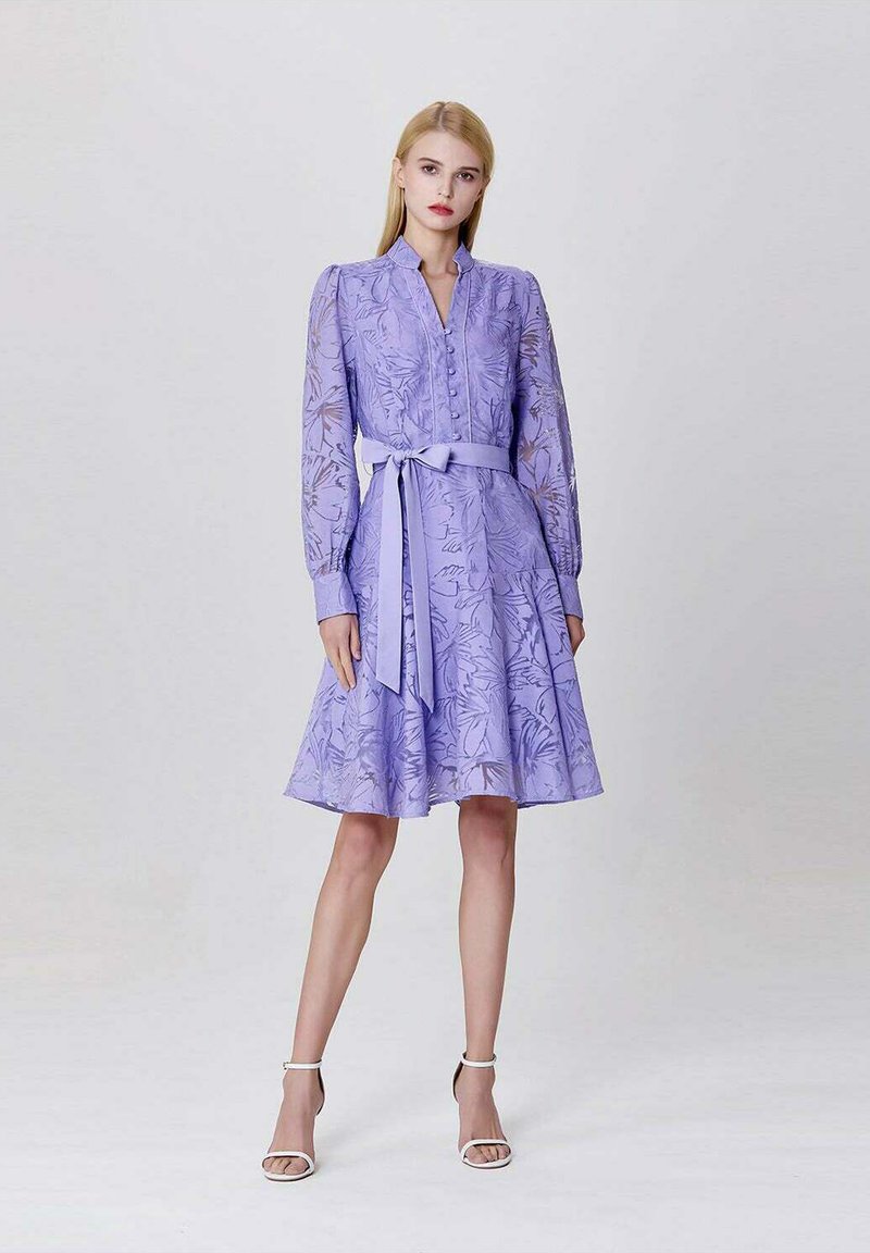 Robe violette à manches longues, motif floral, avec un devant boutonné et une ceinture à la taille. Jupe évasée et arrivant au genou. Associée à des sandales blanches.