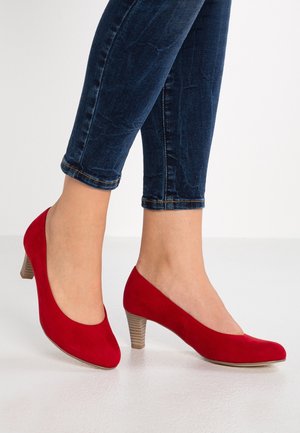 Frau trägt dunkelblaue Skinny-Jeans und rote Wildleder-Pumps mit mittlerem Absatz, steht auf einer weißen Oberfläche vor einem einfarbigen Hintergrund.