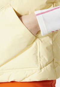 Světle žlutá péřová bunda s boční kapsou, vyrobená z hladkého materiálu s kontrastním bílým dlouhým rukávem s růžovým prošíváním.