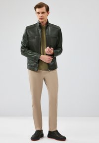 Giacca di pelle verde scuro con colletto alto, due tasche pettorali con zip, pantaloni beige e sneakers nere con dettagli arancioni.