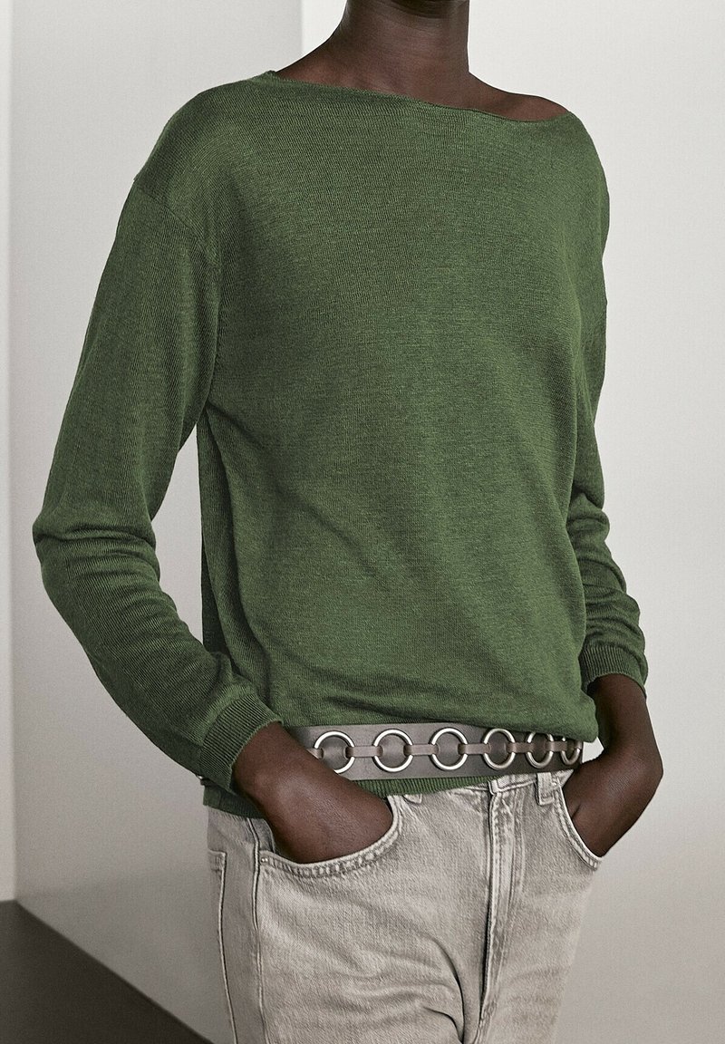 Personne portant un pull vert olive rentré dans un jean beige clair, les mains dans les poches, avec une ceinture à maillons en argent visible.