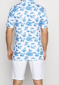 Krátký rukáv košile v bílé barvě s potiskem modrého pastorálního výjevu, zobrazující zvířata a postavy. Kombinujte s bílými šortkami pro koordinovaný vzhled.