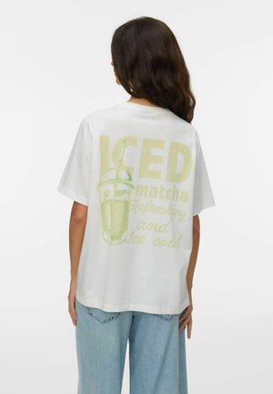 Kvinde med langt mørkt hår iført hvid T-shirt med lysegrøn iset matcha-drik grafik og tekst, kombineret med lyseblå jeans.