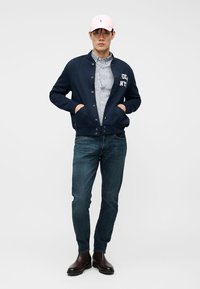 Ung mann iført rosa cap, marineblå knappet jakke, blå jeans, brune støvler og en stripete skjorte mot en ensfarget bakgrunn.