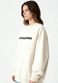 Sweat-shirt beige clair à manches longues, col rond, avec un texte graphique noir "WATCH & DRAMA" sur le devant. Matière douce en mélange de coton.