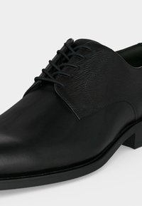 Chaussure habillée en cuir noir avec lacets, panneau texturé près de la cheville et un petit talon sur fond uni.