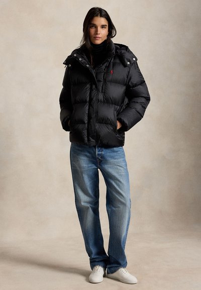 Mujer de pie con las manos en los bolsillos, lleva una chaqueta acolchada negra, vaqueros azules, zapatillas blancas y un cuello vuelto negro sobre un fondo liso.