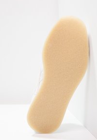 Sula av vit sko med beige texturerad gummiyttersula, lutad upprätt mot vit vägg och golv i bakgrunden.