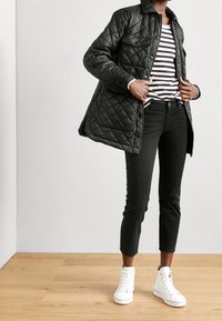 Veste extérieure matelassée noire sur un haut rayé, associée à un pantalon skinny noir et des baskets montantes blanches. Fond en parquet.