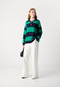 Grün- und marineblau gestreifter oversized Pullover mit kragenförmigem Ausschnitt und drei Knöpfen, kombiniert mit weit geschnittenen weißen Hosen und einer schwarzen Tasche.