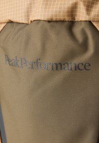 A felső részen bézs kockás anyag, az alján pedig texturált olívazöld anyag található. Az alsó szakaszon szürke "Peak Performance" logó látható.
