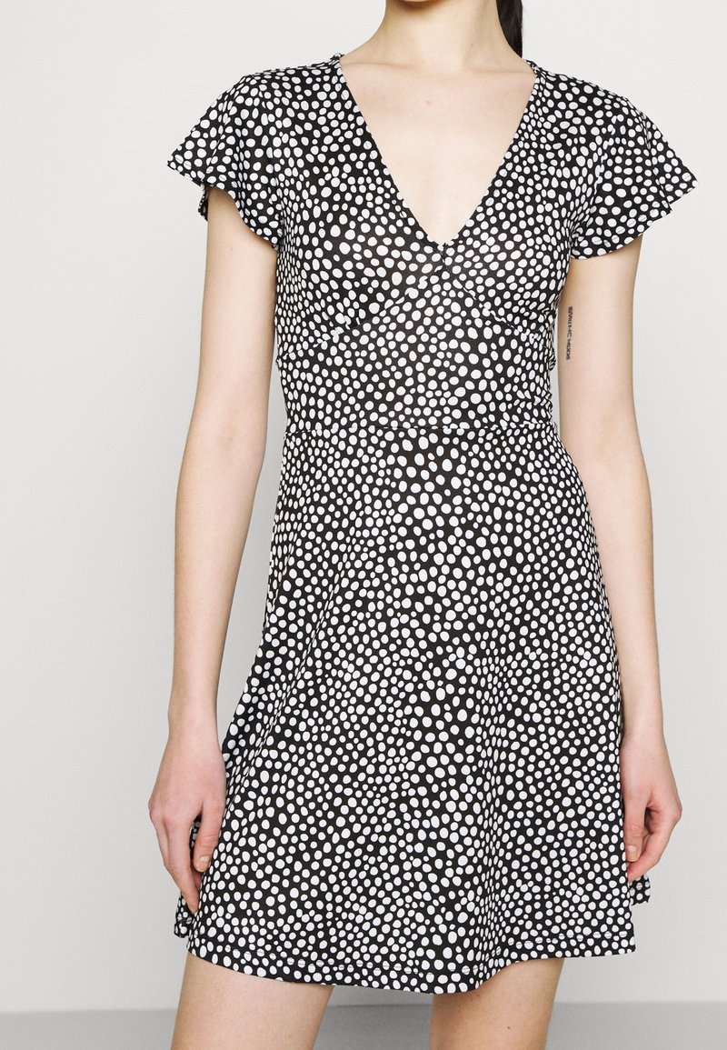 Personne portant une robe noire à manches courtes, à pois blancs et à encolure en V, debout devant un fond clair et uni.