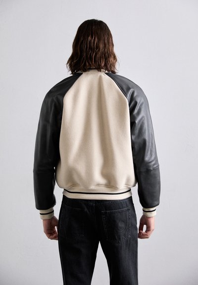 Kremna in črna bombažna jakna z usnjenimi rokavi, teksturirano telo, rebrasti manšeti in dno, oblečena s črnimi hlačami, prikazana od zadaj.
