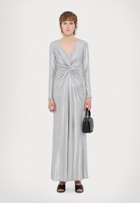 Ασημί μεταλλικό μακρύ φόρεμα με μακριά μανίκια και βαθύ V-ντεκολτέ. Διαθέτει σ�τριμμένο μπροστινό σχέδιο και ρέουσα φούστα. Ζευγάρι μαύρες σάντες.