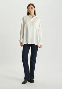 Chemise blanche à manches longues et boutonnée, avec une coupe décontractée, associée à un jean en denim foncé et des chaussures plates noires. Tissu lisse, col standard.