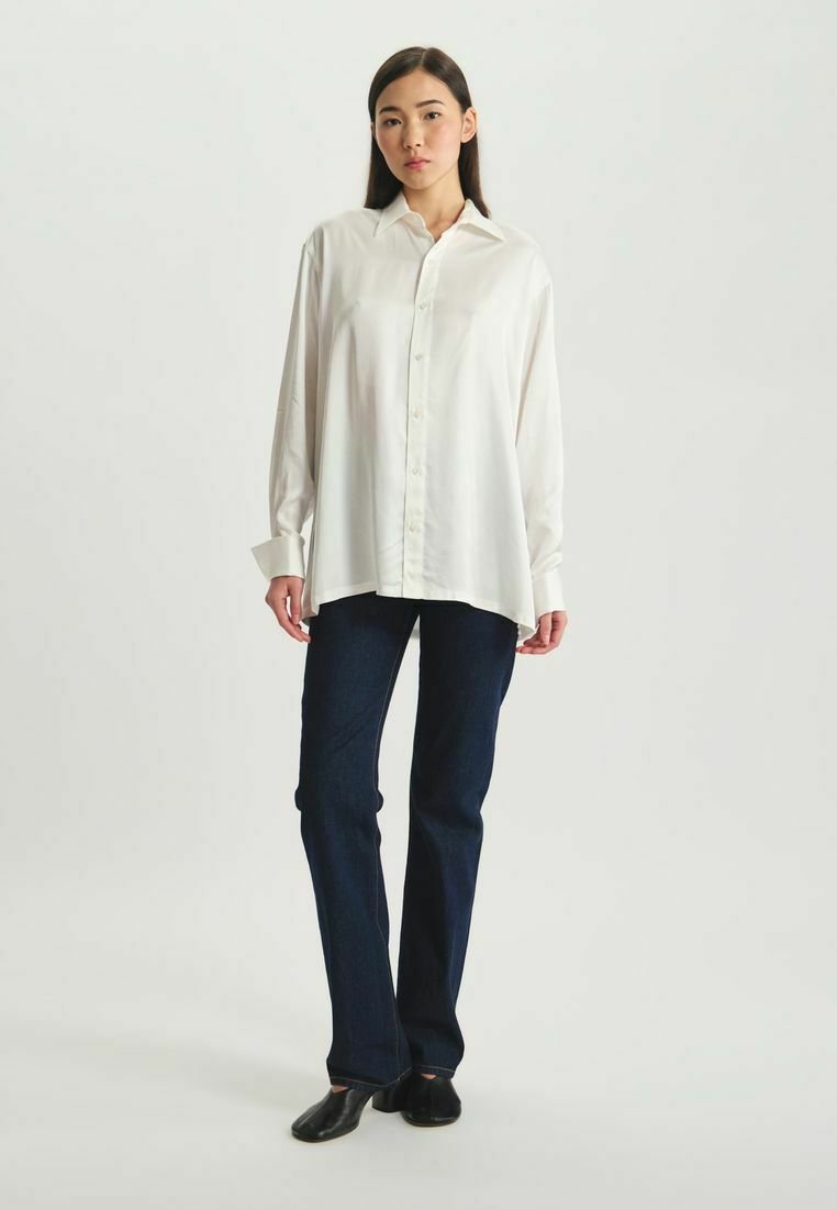 Chemise blanche à manches longues et boutonnée, avec une coupe décontractée, associée à un jean en denim foncé et des chaussures plates noires. Tissu lisse, col standard.