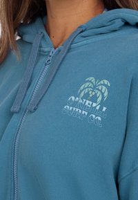 Blauwe ritssluiting hoodie van zachte stof, met een palmboomlogo en de tekst "O'Neill Surf Co." in het wit. Inclusief trekkoord bij de capuchon.