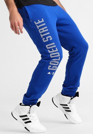Personne portant un pantalon de survêtement bleu Golden State et des baskets blanches Adidas, debout avec les jambes fléchies devant un fond uni.