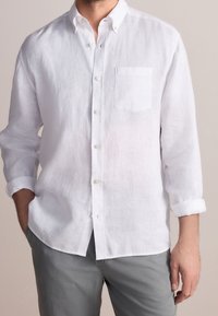 Homme portant une chemise blanche à manches longues avec poche avant et un pantalon gris, une main dans la poche, manches retroussées.
