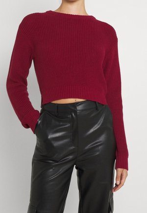Femme portant un pull court en maille rouge foncé et un pantalon en cuir noir taille haute, avec une main dans la poche, sur un fond uni.