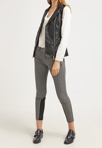 Schwarze Weste aus Kunstleder über einem weißen Langarmshirt, kombiniert mit grauen, karrierten Slim-Fit-Hosen mit schwarzen Akzenten und schwarzen Schnallenschuhen.