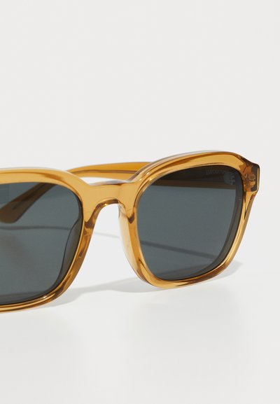 Lunettes de soleil avec monture en plastique ambre translucide, verres teintés gris, forme carrée et design simple. Logo notable sur la branche.