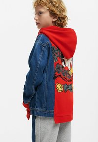 Denim jacket with red hood. Features a graphic print of a character with flames and the text "MICKEY." Textured fabric and contrasting colors.