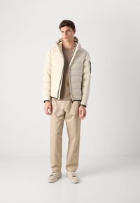 Jonge man staand, gekleed in een beige pufferjack over een taupe shirt, beige broek en lichtbeige sneakers tegen een effen lichte achtergrond.